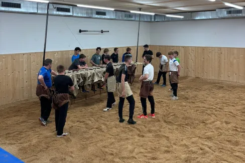 Innenaufnahme einer grossen Halle mit Holzdecke und Holzverkleidung an den Wänden. Mehrere junge Männer stehen in Arbeitskleidung auf einem mit Sägemehl bedeckten Boden und arbeiten gemeinsam an Holzbänken. Die Szene vermittelt eine handwerkliche Ausbildungs- oder Trainingssituation in einer Werk- oder Übungshalle.