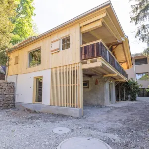 einblicke in ein holzbauprojekt in gunten: die aussenansicht zeigt ein haus mit heller holzfassade und balkon, eingebettet zwischen bäumen und einem steinmäuerchen. im inneren prägen helle holzdecken und sichtbare balken, grosse fenster und warme holzoberflächen die räume. zu sehen sind ein grosszügiger wohnbereich mit zugang zum balkon und ausblick ins grüne, eine holzküche mit arbeitsinsel sowie ein badezimmer mit rundem spiegel und aufgesetztem waschbecken. helles ziegelmauerwerk und sorgfältig erhaltene