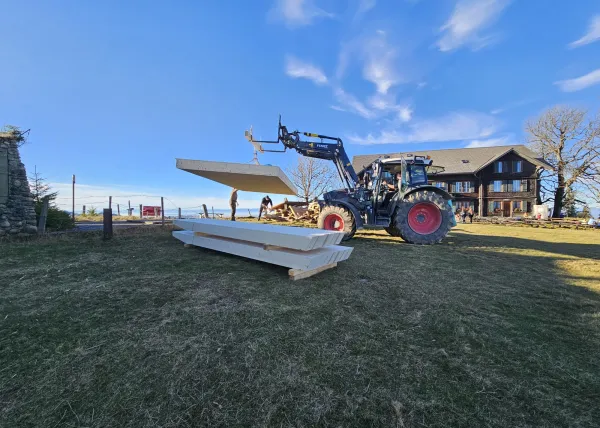 Montagearbeiten an einer Holzplattform vor einem traditionellen Gebäude auf einer Anhöhe. Ein Traktor mit Hebevorrichtung hebt einen langen Holzbalken an, während zwei Mitarbeitende der Zimmerei Hirschi AG den Balken ausrichten.