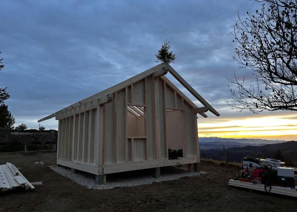 Montagearbeiten an einer Holzplattform vor einem traditionellen Gebäude auf einer Anhöhe. Ein Traktor mit Hebevorrichtung hebt einen langen Holzbalken an, während zwei Mitarbeitende der Zimmerei Hirschi AG den Balken ausrichten.