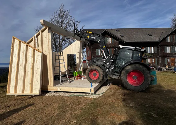 Montagearbeiten an einer Holzplattform vor einem traditionellen Gebäude auf einer Anhöhe. Ein Traktor mit Hebevorrichtung hebt einen langen Holzbalken an, während zwei Mitarbeitende der Zimmerei Hirschi AG den Balken ausrichten.