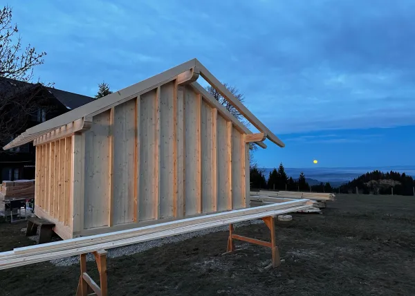 Montagearbeiten an einer Holzplattform vor einem traditionellen Gebäude auf einer Anhöhe. Ein Traktor mit Hebevorrichtung hebt einen langen Holzbalken an, während zwei Mitarbeitende der Zimmerei Hirschi AG den Balken ausrichten.