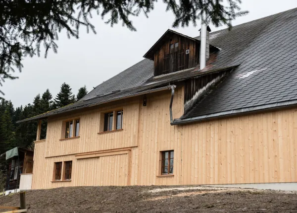 Ausschnitt eines überdachten Balkons in traditioneller Holzbauweise mit hellem Holzgeländer und Holzfassade. Sichtbar sind Fensteröffnungen und das schützende Dach mit Holzbalken. Im Hintergrund öffnet sich der Blick auf eine hügelige Landschaft mit Wäldern und Bergen.
