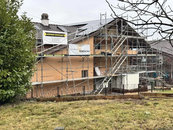 Holzhaus im Bau mit eingerüsteter Fassade und Baustellenbanner der Zimmerei Hirschi unter blauem Himmel.