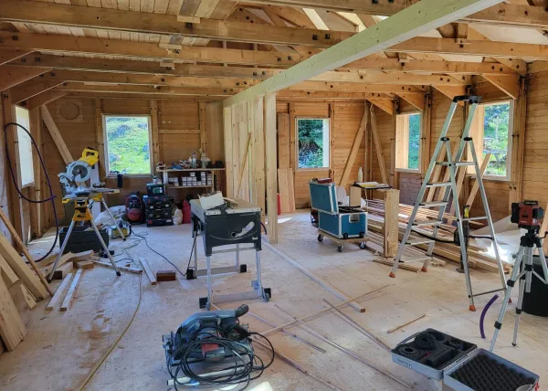 Holzhaus im Bau mit natürlicher Holzfassade inmitten einer Waldlandschaft. Das eingeschossige Gebäude steht auf einem gepflegten Grundstück mit Wiese und Zufahrtsweg. Die Fensteröffnungen sind bereits erstellt, während der Innenausbau noch im Gange ist. Im Hintergrund befinden sich weitere Holzbauten sowie hohe Nadelbäume, die das Gebäude umgeben. Die Aufnahme zeigt die harmonische Einbindung des Holzbaus in die natürliche Umgebung.